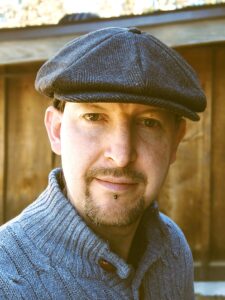 portrait of a man with a goatee in a newsboy cap and gray sweater posing in front of a rich wood wall background