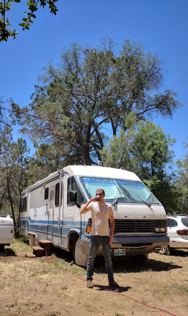 man drinking coffee in front of a vintage RV in the woods